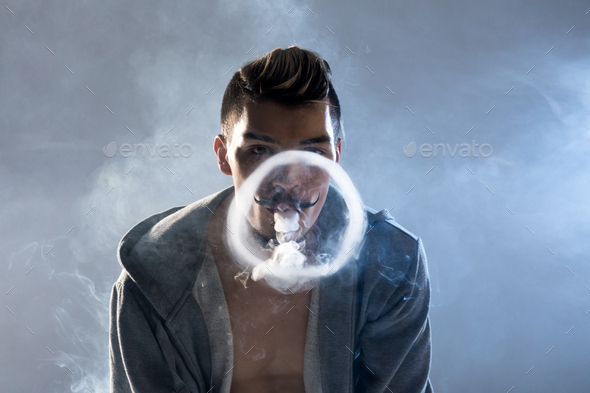 Young bearded man vaping and making smoke circle looking at camera isolated on grey