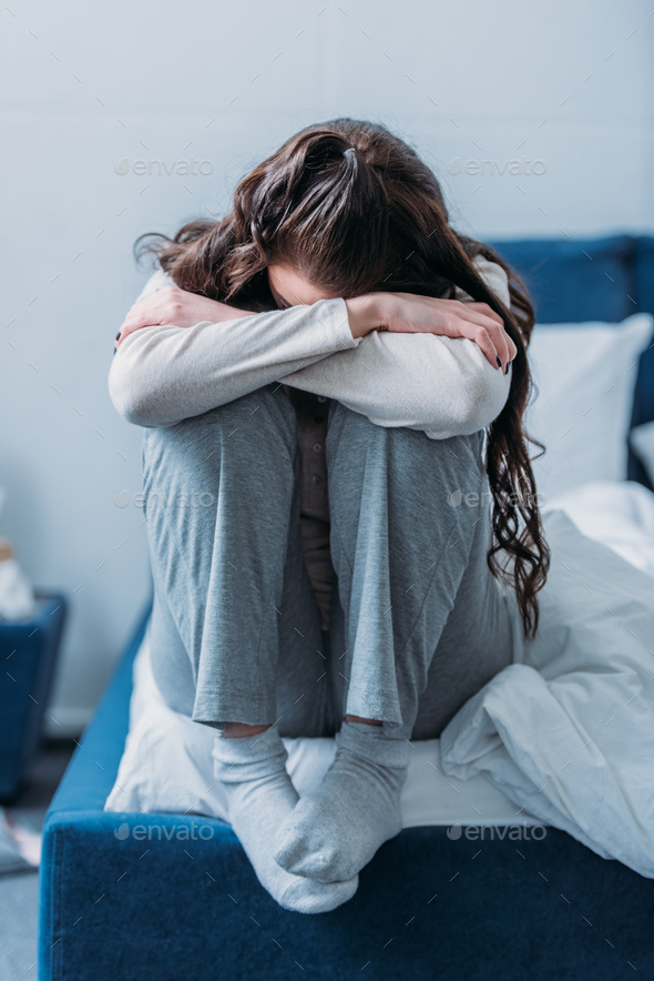 depressed woman with head down hugging knees and sitting on bed at home