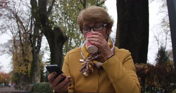 Adult Woman Sitting on a Park Bench, Looking at Her Mobile Phone Drink Coffee. A Woman Using a alt
