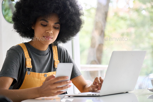 Black hipster girl sitting in cafe using phone and laptop shopping online.