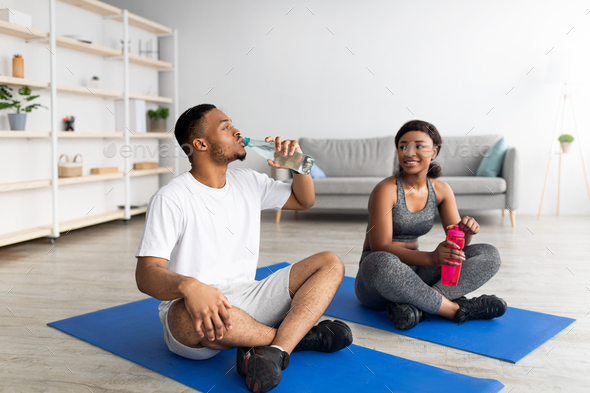 Athletic black couple in sportswear sitting on mats, drinking water from bottles after domestic Stock by