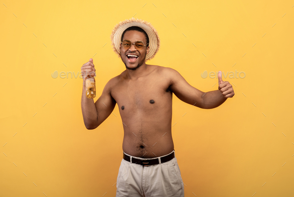 Handsome black guy with naked torso holding bottle of beer and showing thumb up on yellow studio