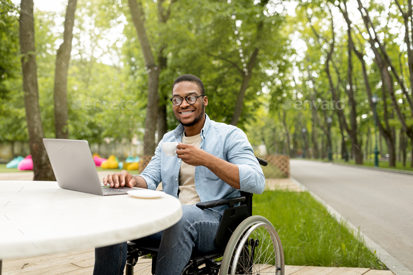 Happy handicapped black man in wheelchair using laptop for online job at cafe in city park