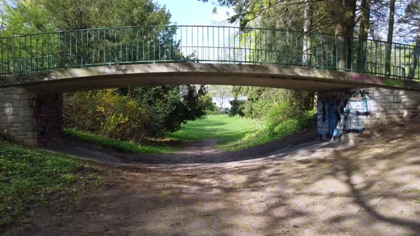 Along a path under a footbridge through into a park. Unbelievable aerial view flight fly forwards dr alt
