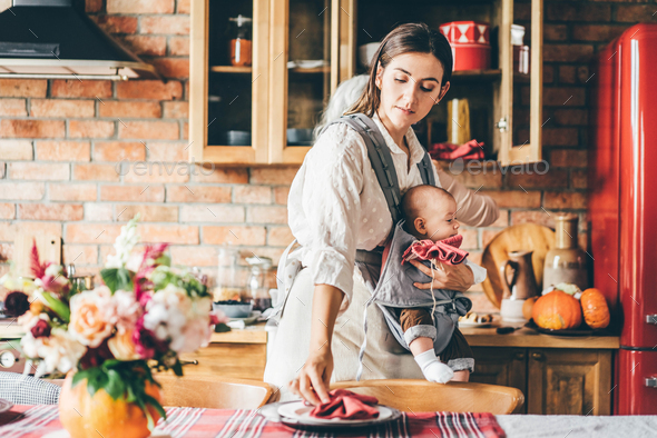 woman hair holds baby in sling bag and helps aged mother serving large table with plates