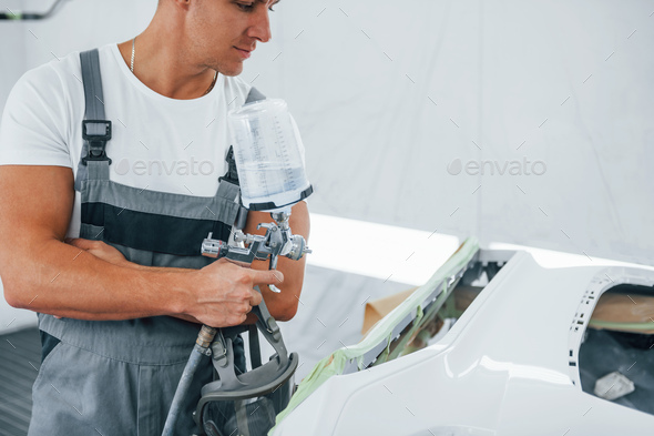 With painting gun. Caucasian automobile repairman in uniform works in garage