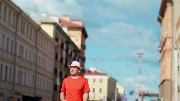 Young Male Tourist in White Hat Walks Along City Street Camera Tracking alt