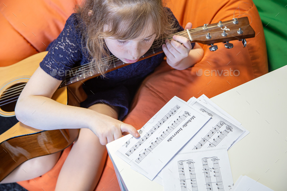 A little girl learns to play the guitar by sheet music.