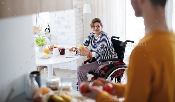 Disabled mature woman in wheelchair sitting at the table indoors at home, talking to friend - Stock Photo - Images