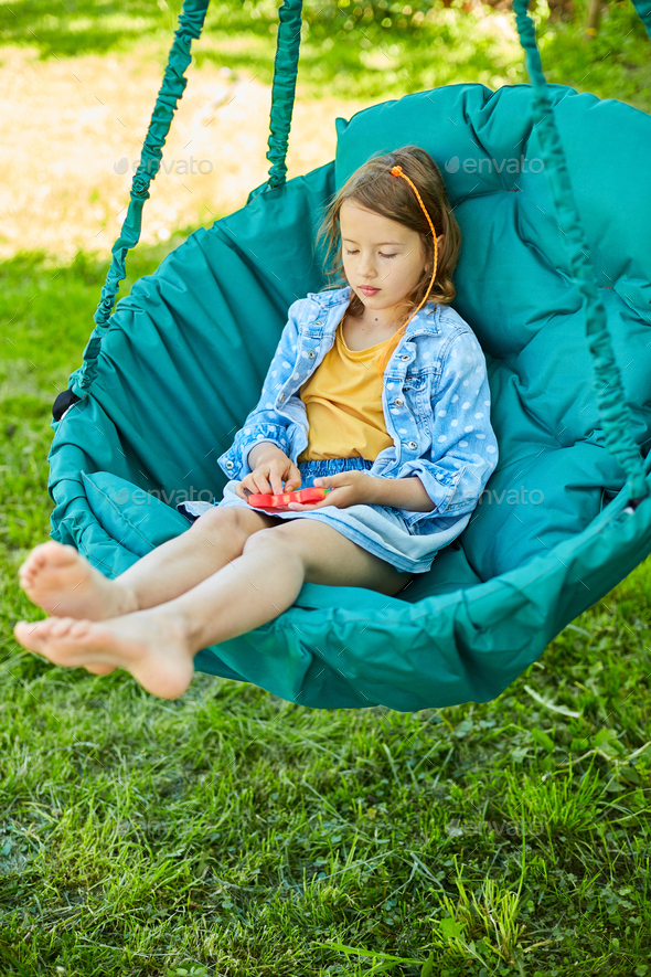 A little girl the on hanging chair outdoors play pop it, kid hands playing with colorful pop It - Stock Photo - Images