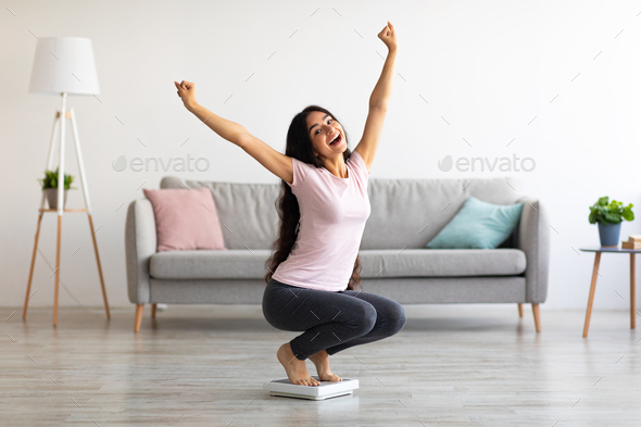Excited Indian woman sitting on scales, raising hands up, happy with result of her weight loss