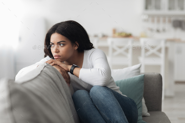 Lonely depressed african american woman sitting on couch with sad face expression and looking away