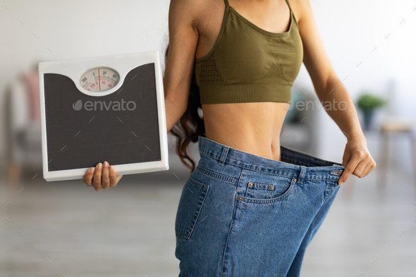 Closeup of Indian woman in oversized jeans holding scales, showing results of weight loss program or