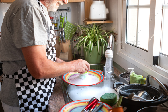 Senior man with white beard concentrated while washing dishes at home wearing apron. Bright light - Stock Photo - Images