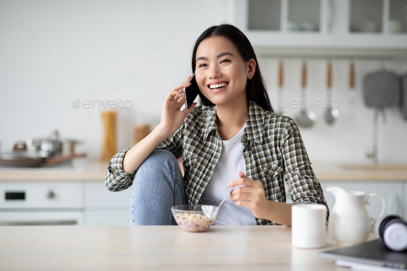 Young asian lady having breakfast and talking on phone - Stock Photo - Images