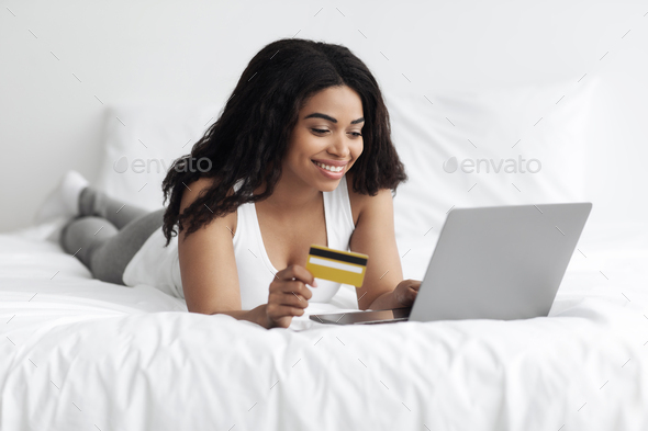 Online payment. Excited black lady using laptop and debit credit card, lying on bed and shopping
