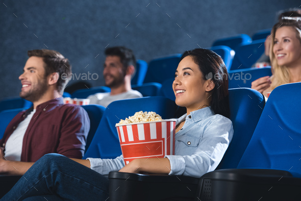 smiling asian woman with popcorn watching movie in cinema alone