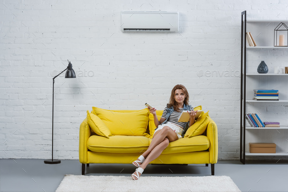 focused young woman with book sitting on sofa and pointing at air conditioner with remote control