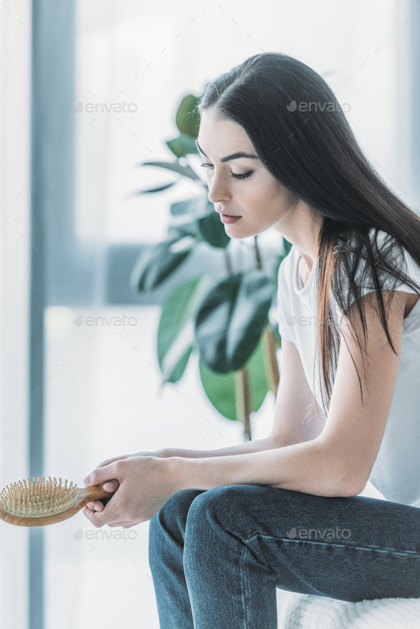 side view of young sad woman holding hairbrush while sitting indoors, hair loss concept - Stock Photo - Images