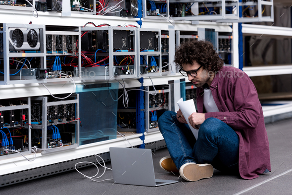 Young computer engineer working while sitting on floor at cryptocurrency mining farm