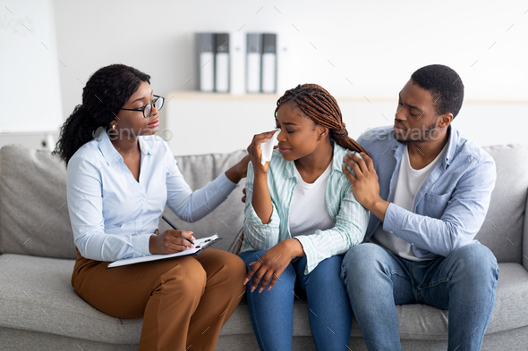 Millennial black woman crying at psychologist\'s office, loving husband supporting her with