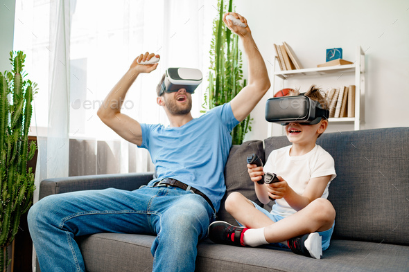 Dad and son sitting on sofa playing games with VR glasses Stock Photo by FabrikaPhoto