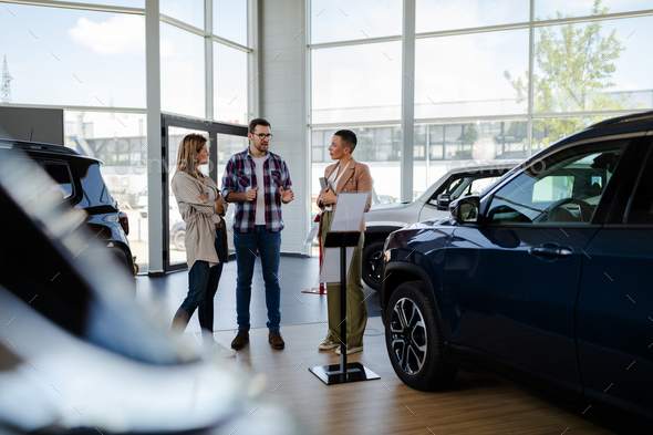 Young female sales agent helping adult couple to choose a new car in modern car showroom.