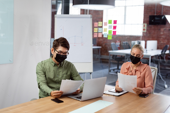 Diverse male and female colleague in face masks sitting at table with laptop reading paperwork - Stock Photo - Images