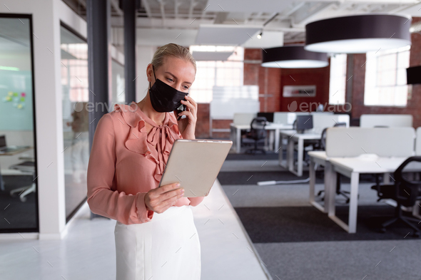 Caucasian businesswoman wearing face mask standing in office using tablet and talking on smartphone - Stock Photo - Images