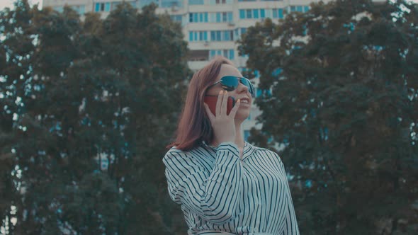 Young Woman in Sunglasses with Mobile Phone on Street in Summertime alt