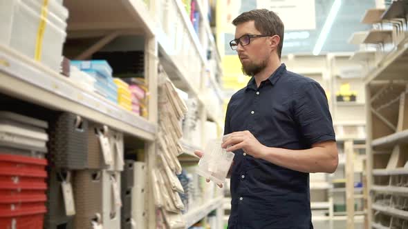 Householder is Comparing Two Plastic Boxes in Supermarket Standing Near Shelves alt