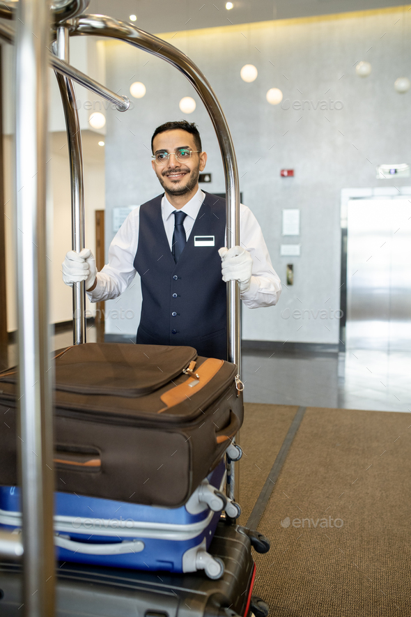 Young porter in uniform pushing cart with baggage - Stock Photo - Images