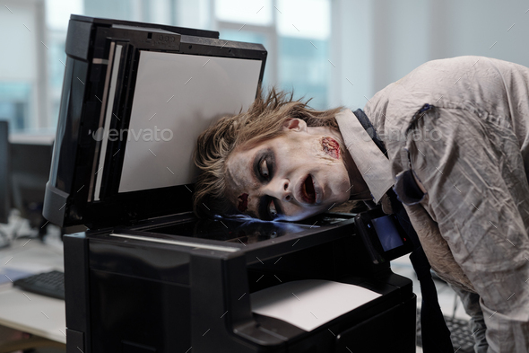 Gloomy man with zombie makeup keeping head on screen of xerox machine