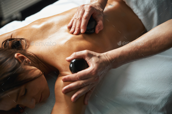 Charming young Caucasian female receiving professional spa procedure with hot mineral stones in spa