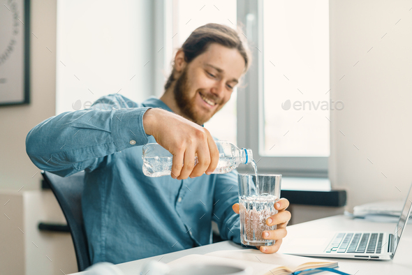 Young happy businessman pouring water into glass at his workplace