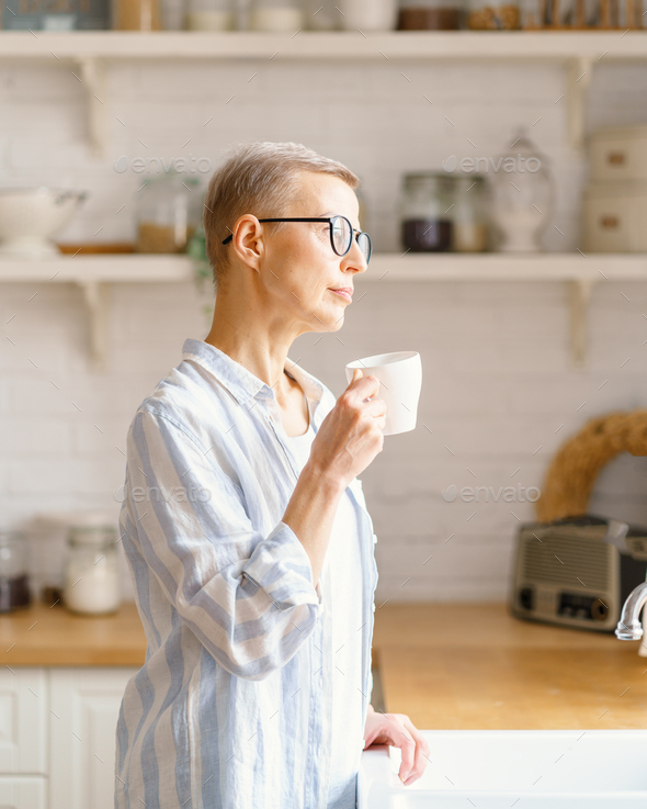 Beautiful senior woman with cup of coffee or rure water looking in window while enjoying new day at - Stock Photo - Images