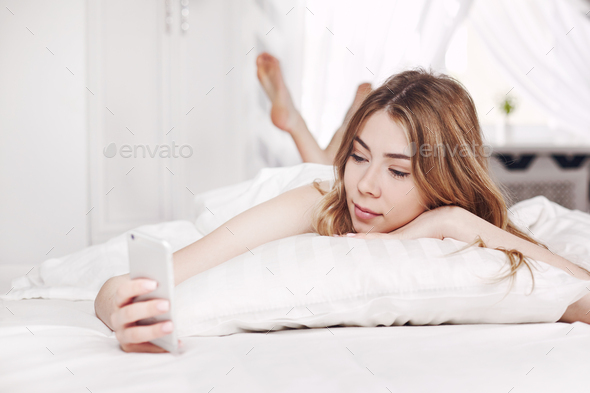 Cute smiling girl looks at the phone, lying on the bed. A girl reads messages on her phone