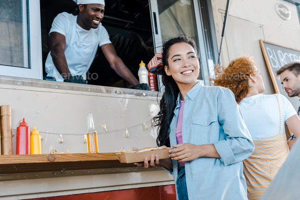 selective focus of cheerful asian girl holding carton plate near customers and chef in food truck