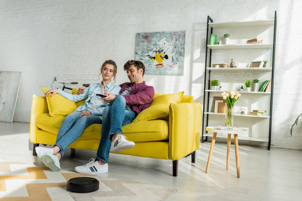 happy man and young woman watching movie while robotic vacuum cleaner washing carpet in living room