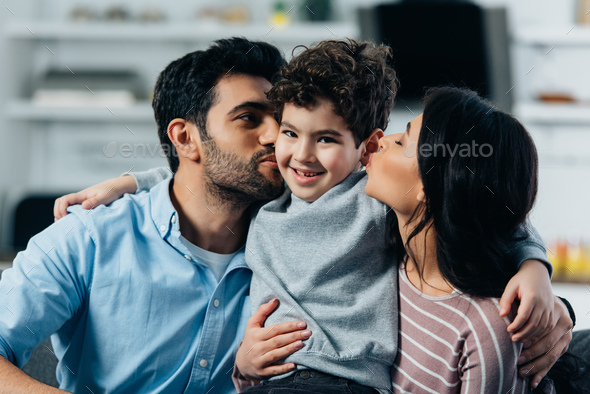 happy latin father and mother kissing cheeks of cute son at home