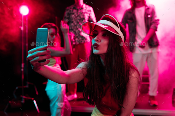 girl pouting lips and taking selfie on smartphone during rave party in nightclub