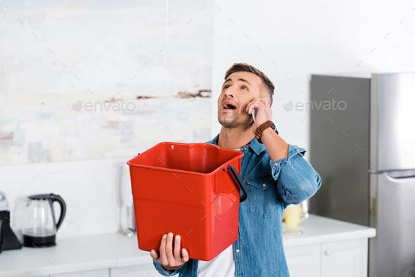 man talking on smartphone while holding plastic bucket and looking at leaking water from ceiling