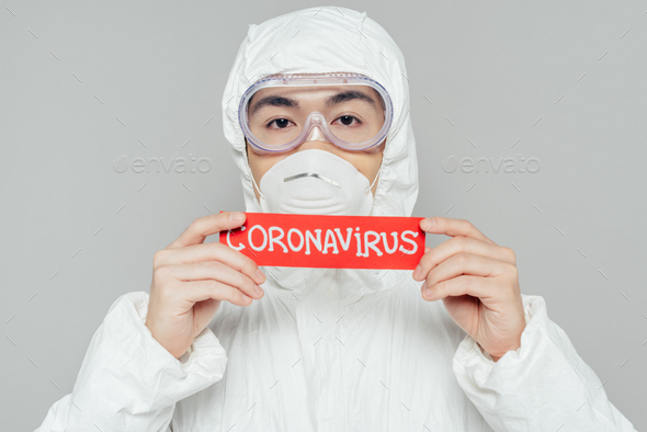 asian epidemiologist in hazmat suit and respirator mask holding warning card with coronavirus