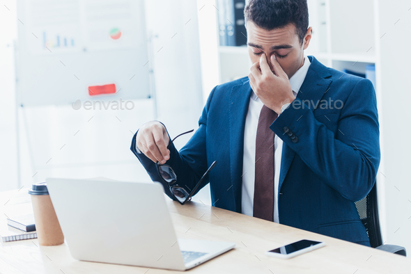 tired young businessman holding eyeglasses and rubbing nose bridge at workplace