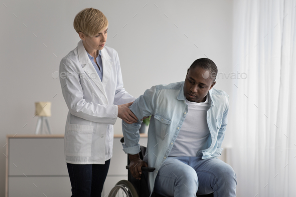 Serious female therapist in white coat looks at mature african american to disabled man - Stock Photo - Images