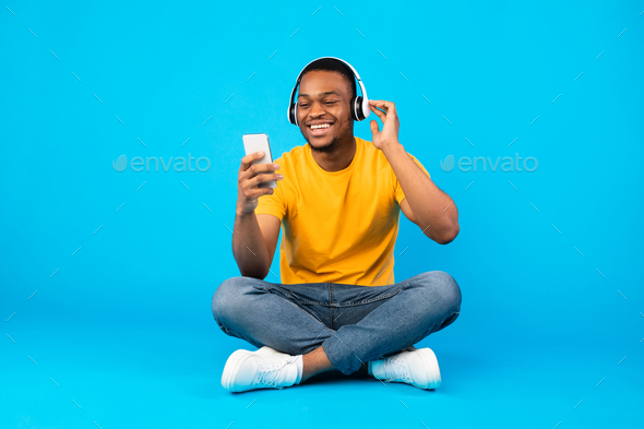 African Guy Listening Music Using Smartphone Wearing Headphones, Blue Background - Stock Photo - Images