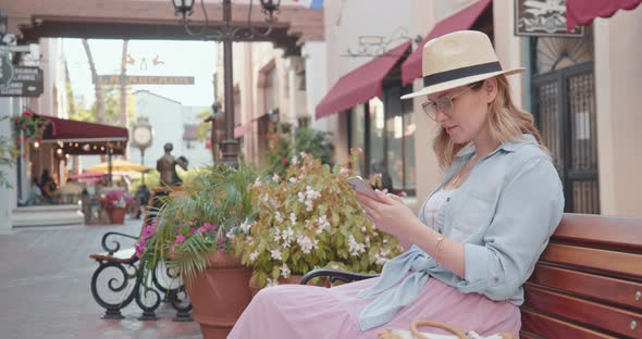 Pretty Woman Is Checking Phone on Summer, Sitting on Bench in Cinematic Old Town alt