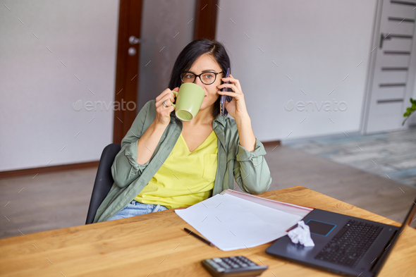 Stylish brunette woman in glasses sitting at wooden table with notepad and having phone call - Stock Photo - Images