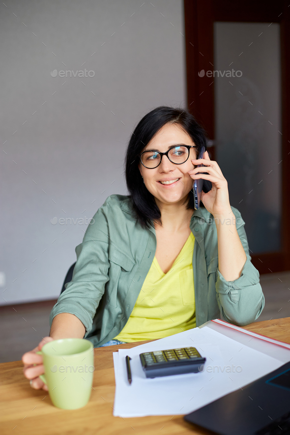 Stylish brunette woman in glasses sitting at wooden table with notepad and having phone call - Stock Photo - Images