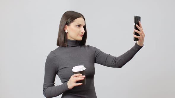 Woman With Coffee Cup In Gray Turtle Neck Dress On A Grey Background Making Selfie On Her Sell Phone